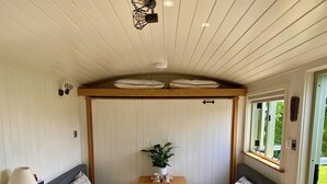 A cozy interior with a wooden table, two seating areas with cushions, a small kitchen area, and a window providing a view of the outdoors. - A quiet and cosy Shepherds Hut, with wood burning fire with scenic views (Conwy)