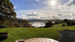 A round wooden table with two patterned coffee mugs, set on a grassy lawn with a scenic view of a lake and hills in the background. - A quiet and cosy Shepherds Hut, with wood burning fire with scenic views (Conwy)