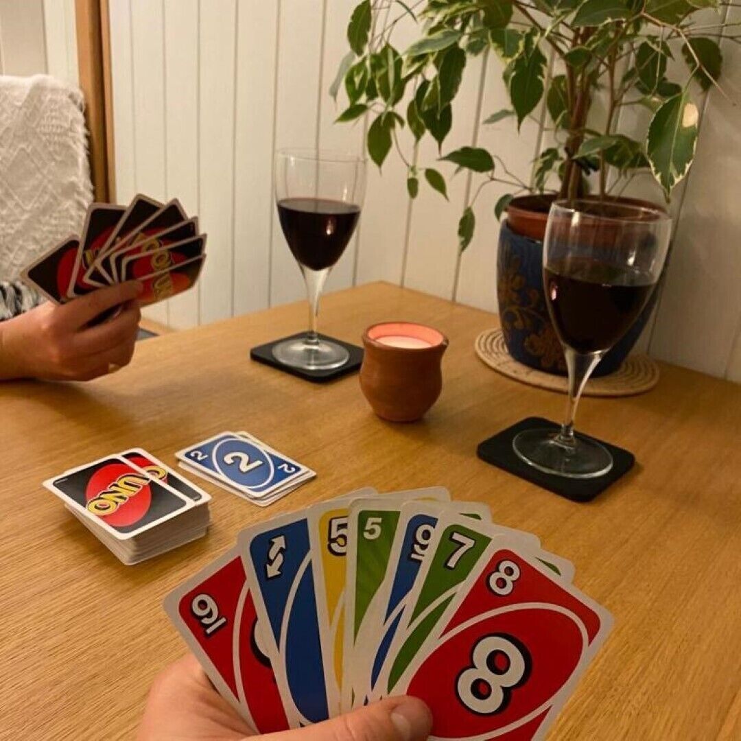 A person holding playing cards at a wooden table with two glasses of red wine, a candle, and a potted plant.