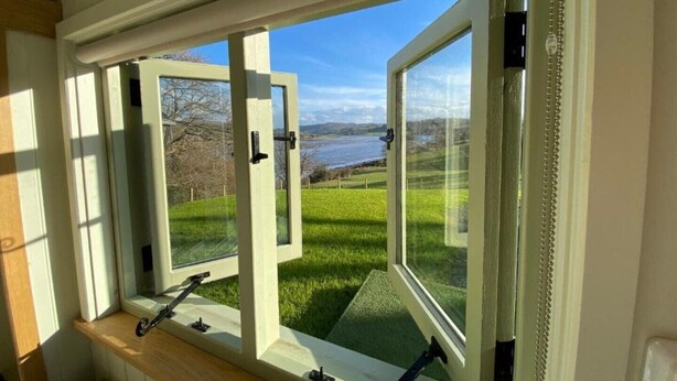 A room with a large window overlooking a grassy area and a body of water. - A quiet and cosy Shepherds Hut, with wood burning fire with scenic views (Conwy)