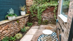 Eine kleine Terrasse im Freien mit einer Steinmauer, einer mit Efeu bewachsenen Backsteinmauer, einem Tisch und Stuhl sowie Blumentöpfen.