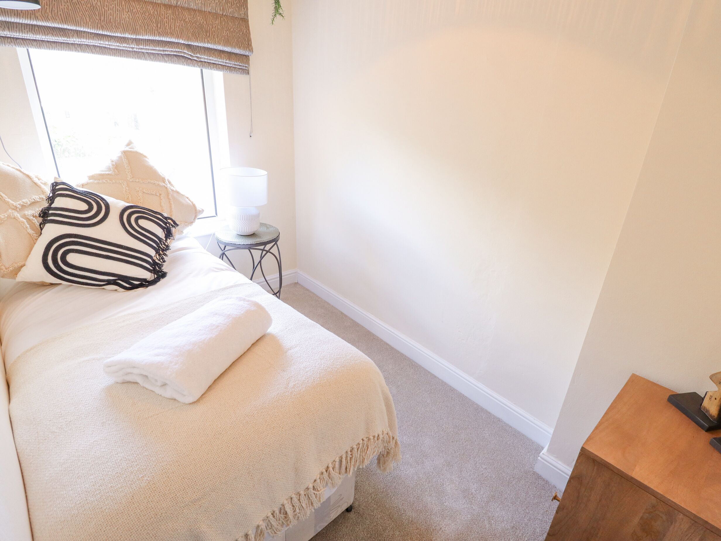Un lit bien fait, avec un oreiller à rayures noires et blanches, une couverture beige et une petite table de chevet surmontée d’une lampe blanche.