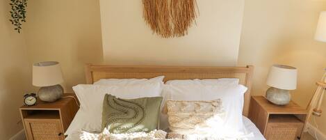Un dormitorio con una cama de madera, dos mesitas de noche con lámparas, un macramé colgado en la pared y una planta suspendida.