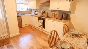 A modern kitchen with wooden cabinets, a built-in oven, a microwave, and a dining area with a table set for two. - Rose Cottage (Brough)