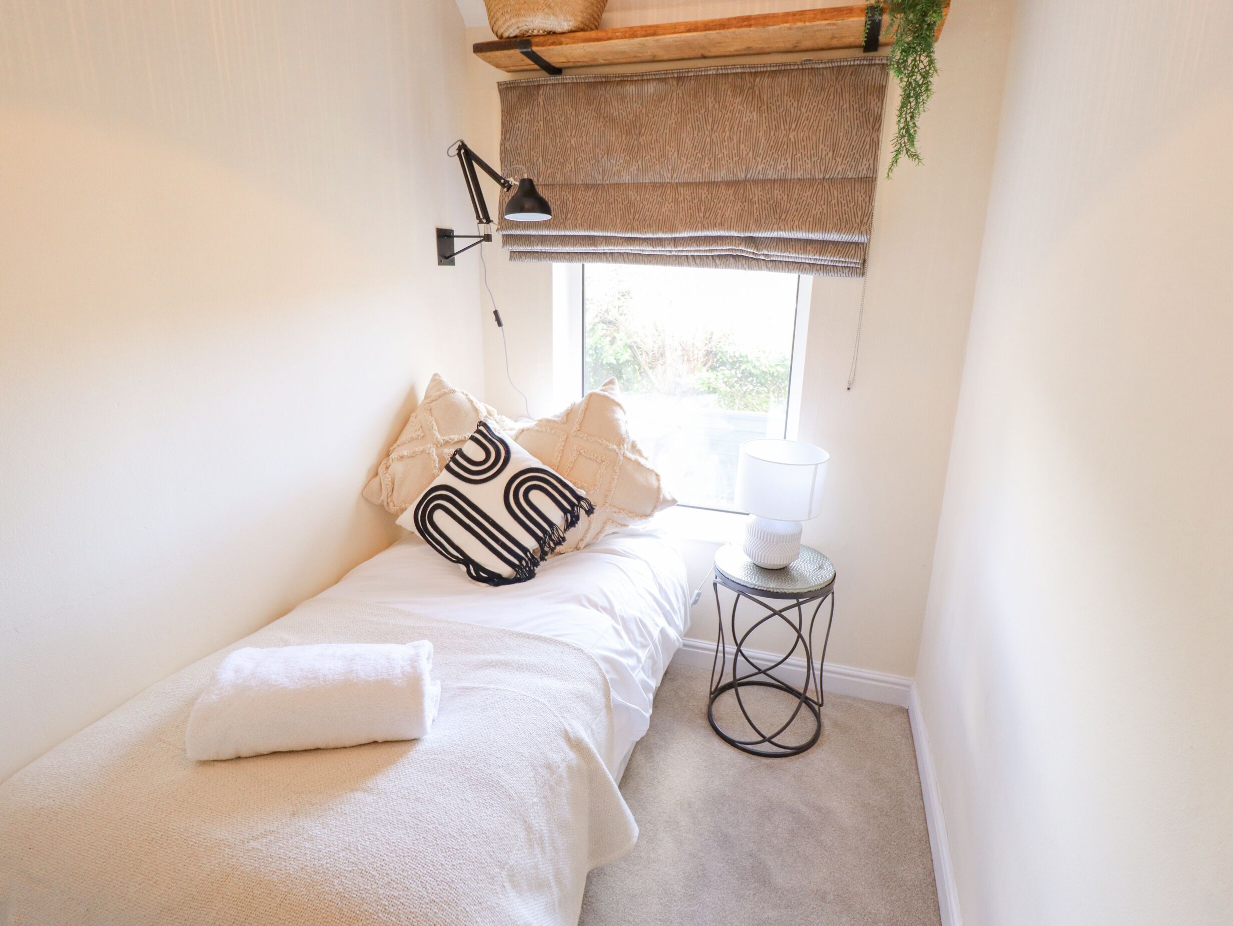 A small, neatly arranged bedroom with a single bed, a bedside lamp, a window with a view, and a small table with a lamp.