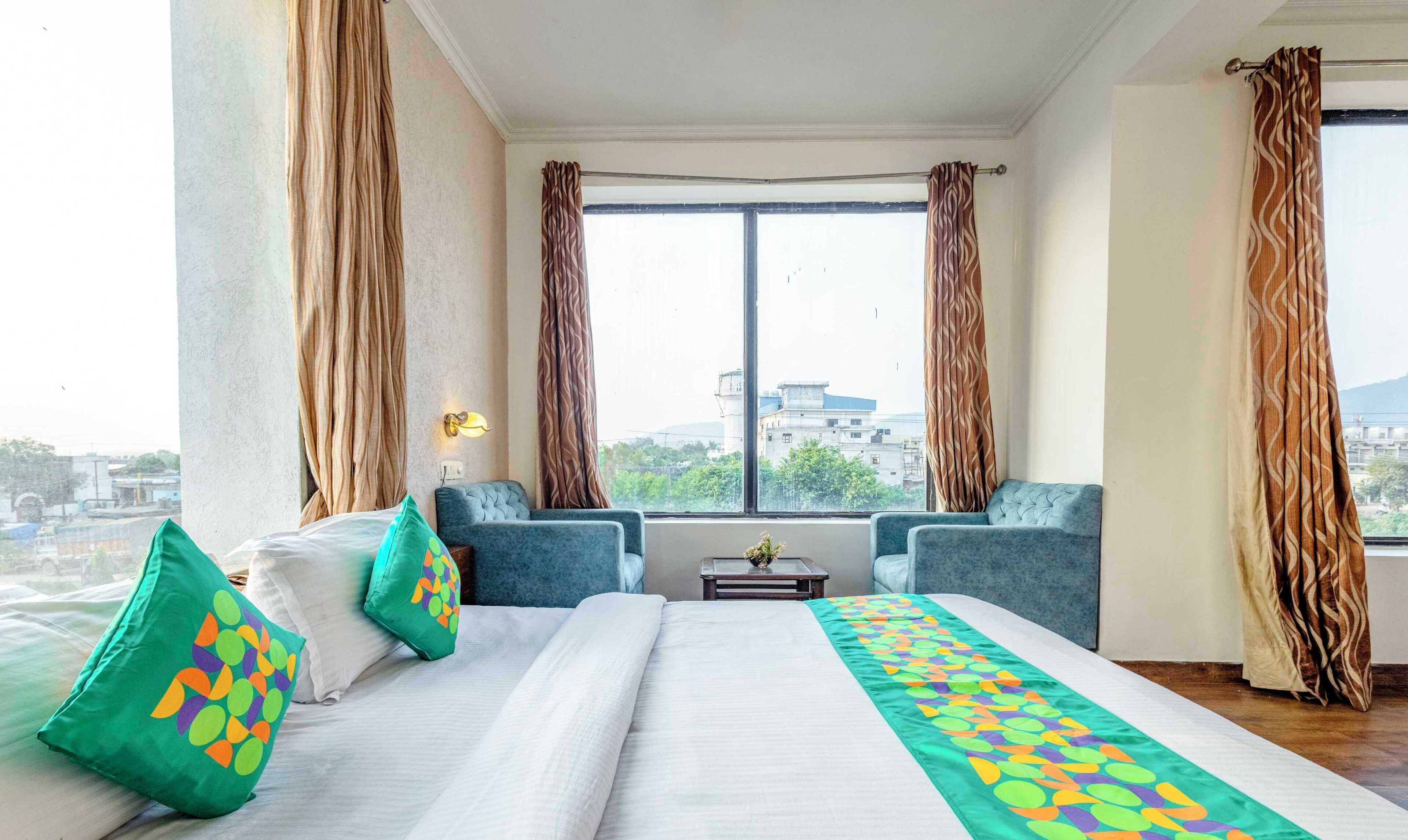 Habitación de hotel con una cama grande, dos sillones, una mesita y vistas a la ciudad a través de amplias ventanas.