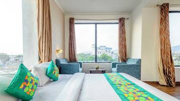 Habitación de hotel con una cama grande, dos sillones, una mesita y vistas a la ciudad a través de amplias ventanas.