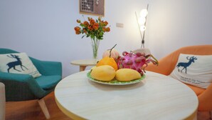 Ein runder Tisch mit einer Fruchtplatte und einer Blumenvase in einem Wohnzimmer mit türkisfarbenen und orangefarbenen Stühlen.