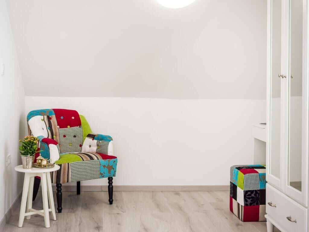 A room with a white wardrobe, a colorful chair with patterned cushions, a small white table with a plant, and a colorful storage box.