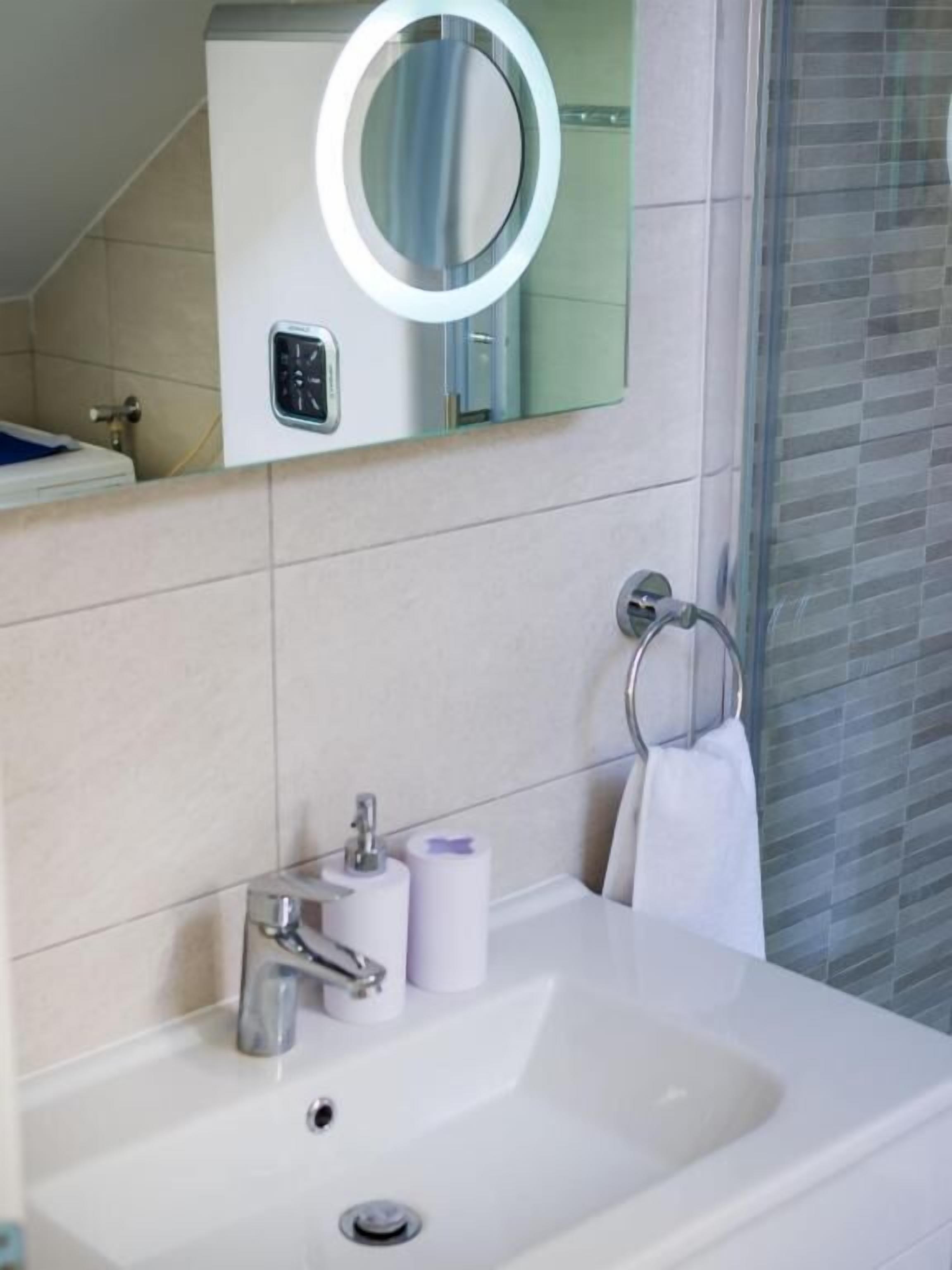 A modern bathroom with a white sink, a mirror with a ring light, a towel rack, and a toilet paper dispenser.