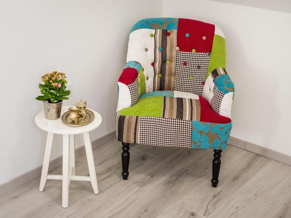 A chair with a patchwork design, a small white table with a plant and a decorative item, and a wooden floor.