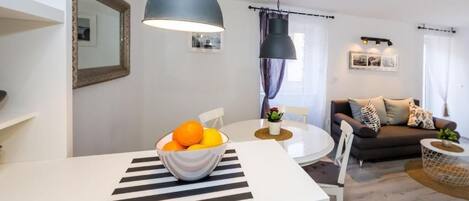 A modern living room with a white dining table, a sofa, and a large window with curtains.