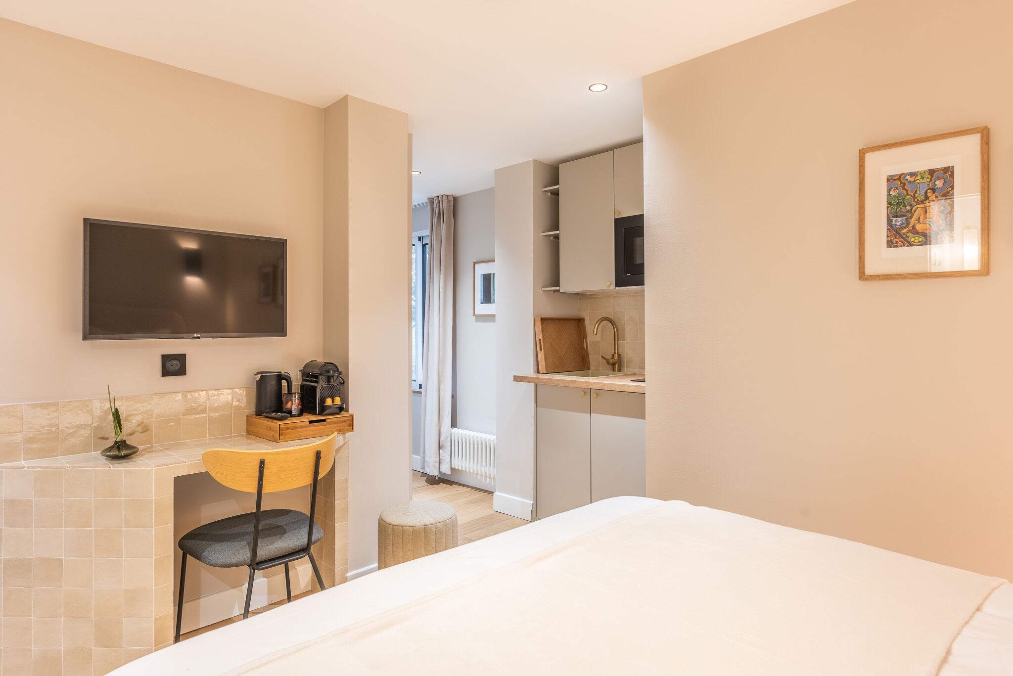 A modern hotel room with a bed, a desk with a chair, a TV, and a kitchenette.