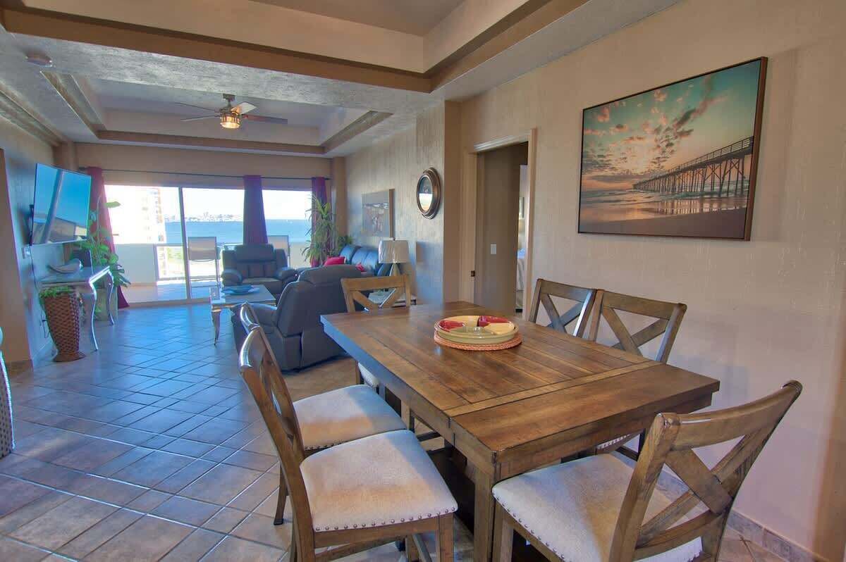 A dining area with a wooden table and chairs, a painting on the wall, and a view of the ocean through large windows.