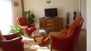 A living room with a television on a wooden cabinet, a sofa, two armchairs, a coffee table, and a chandelier. - Le Fournil (La puisaye)