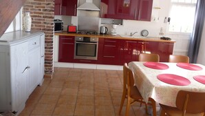 A compact kitchen with red cabinets, a built-in oven, and a dining area with a table set for four. - Le Fournil (La puisaye)