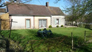 A two-story house with a brick exterior, a green door, and a small front yard with a lawn and a table with chairs. - Le Fournil (La puisaye)