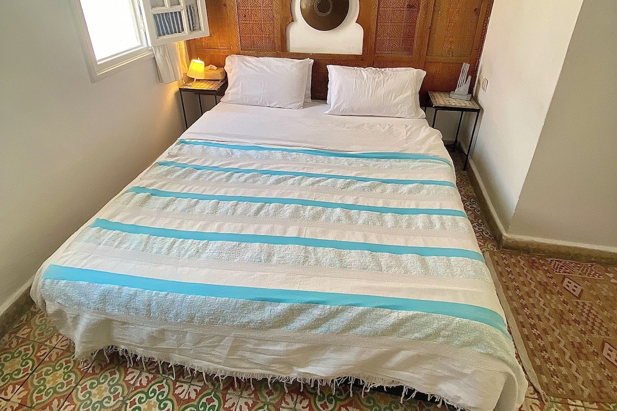 A neatly made bed with a striped bedspread, a wooden headboard with a circular design, a painting above the bed, and patterned floor tiles.