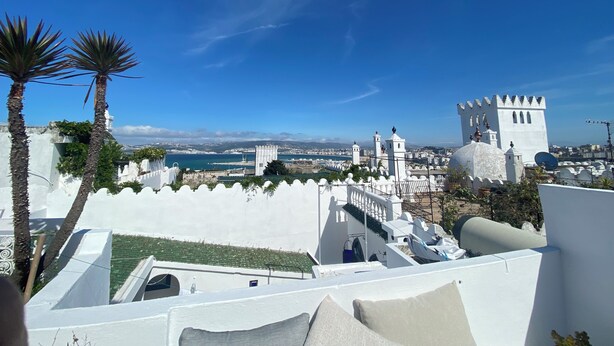 A rooftop terrace with white walls, palm trees, and a view of a coastal town with a castle-like structure. - Bab El Fen  (Tangier)