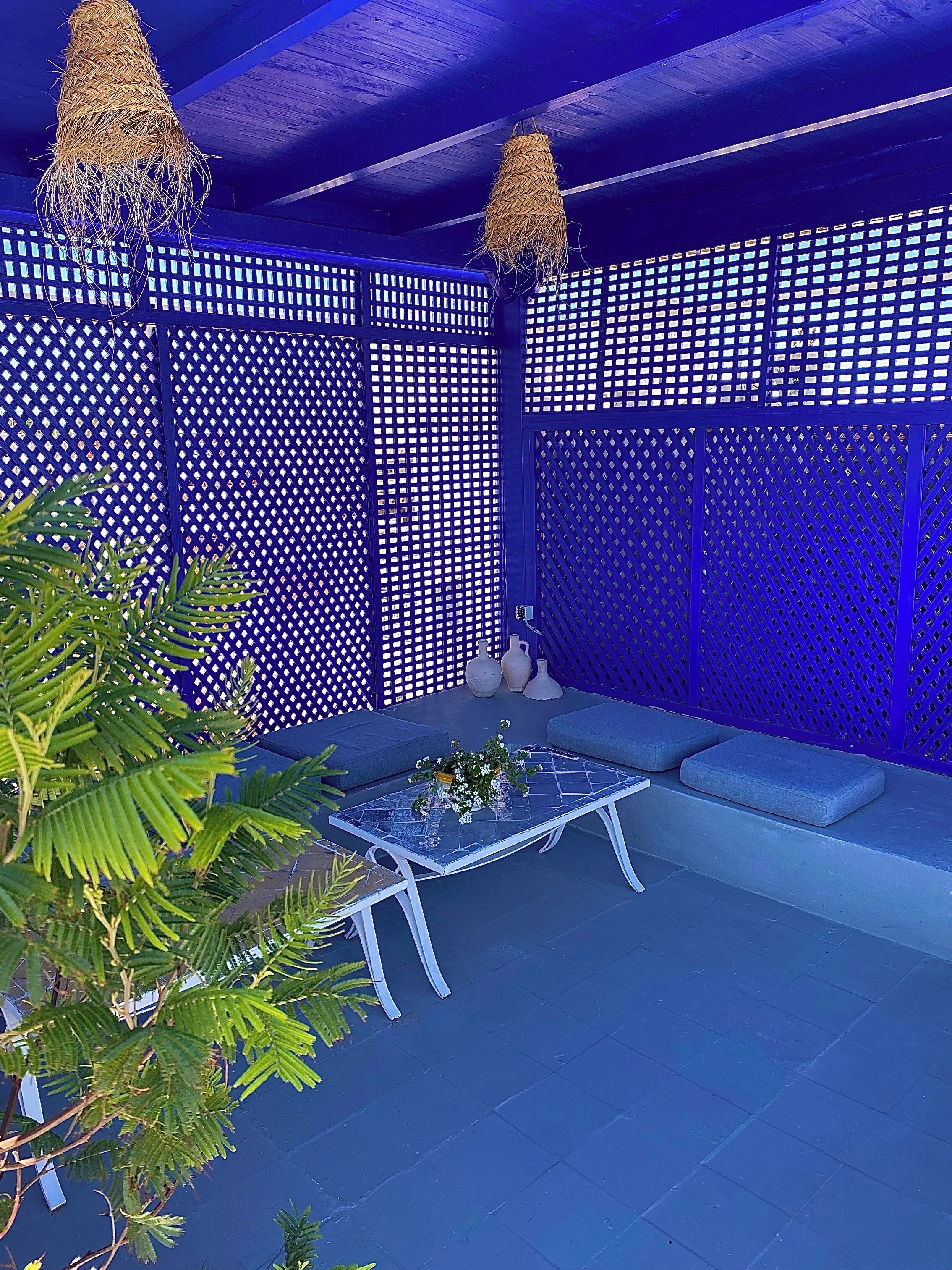 Un espacio al aire libre con decoración azulada, con una mesa y cojines, rodeado por una celosía y plantas en macetas.