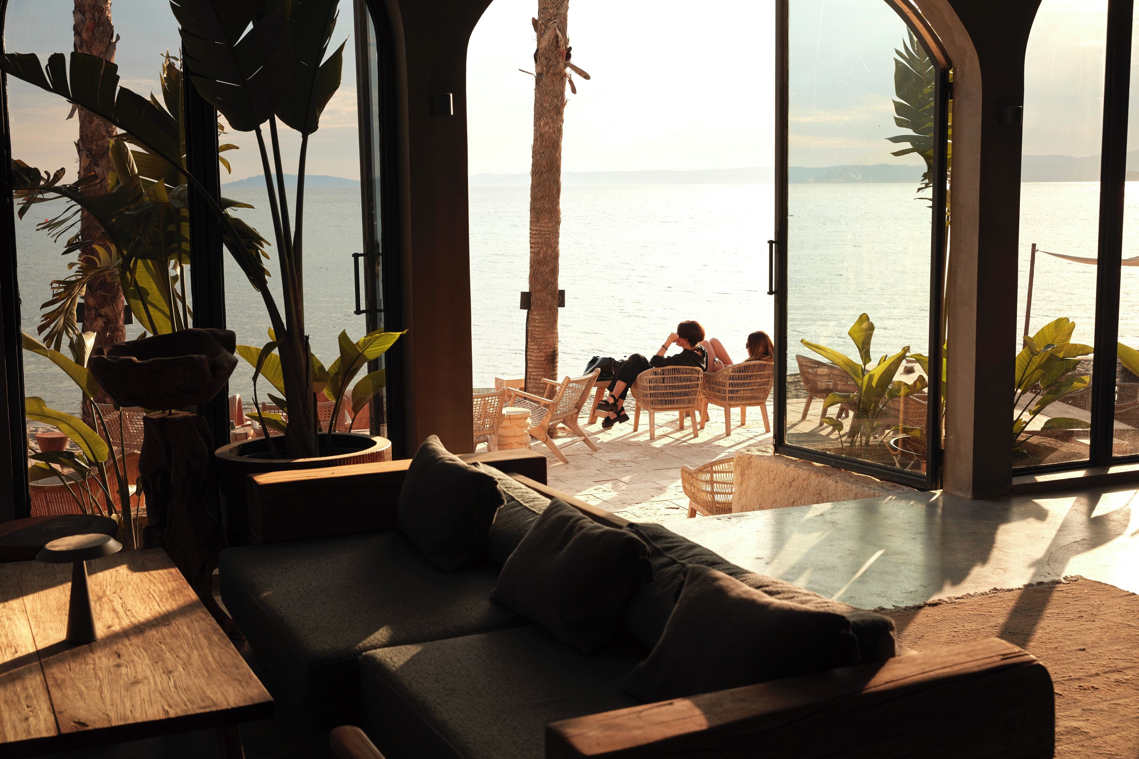 A modern interior with a sofa, wooden tables, and large windows overlooking a beach.