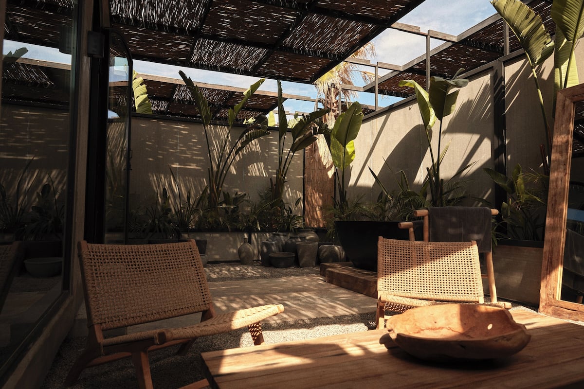 An outdoor seating area with wicker chairs and a wooden table, surrounded by potted plants and a thatched roof structure.
