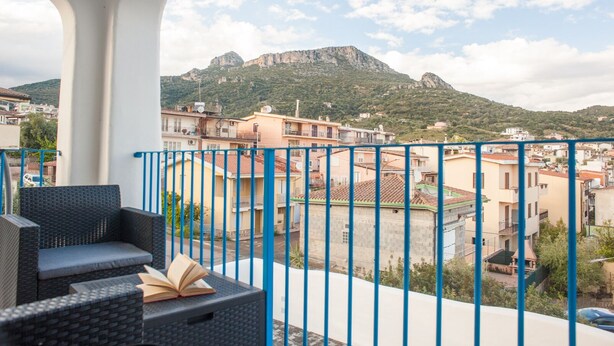 Ein Balkon mit Korbmöbeln und einem Buch, mit Blick auf eine Stadt mit Gebäuden und einem Berg.