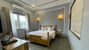 A bedroom with a wooden bed, a bedside table, a window with curtains, and an air conditioning unit. - The Arham Resort (Udaipur)
