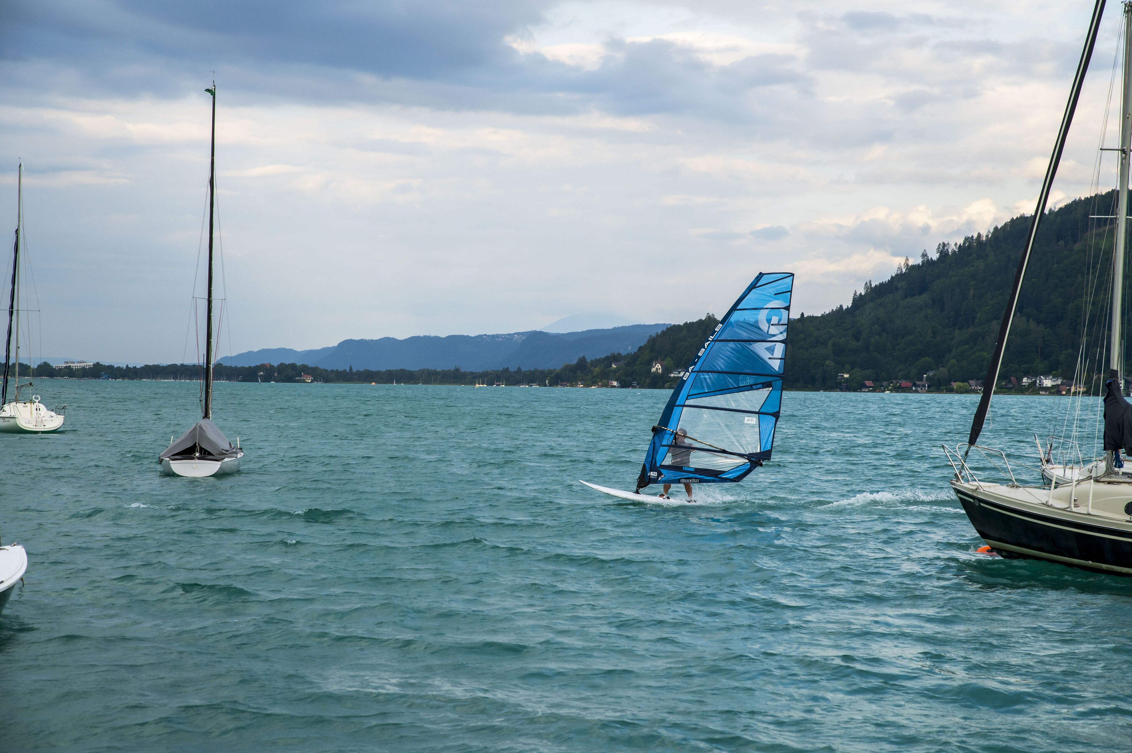 A sailboat with a blue sail on a body of water, surrounded by other boats and a mountainous landscape in the background.