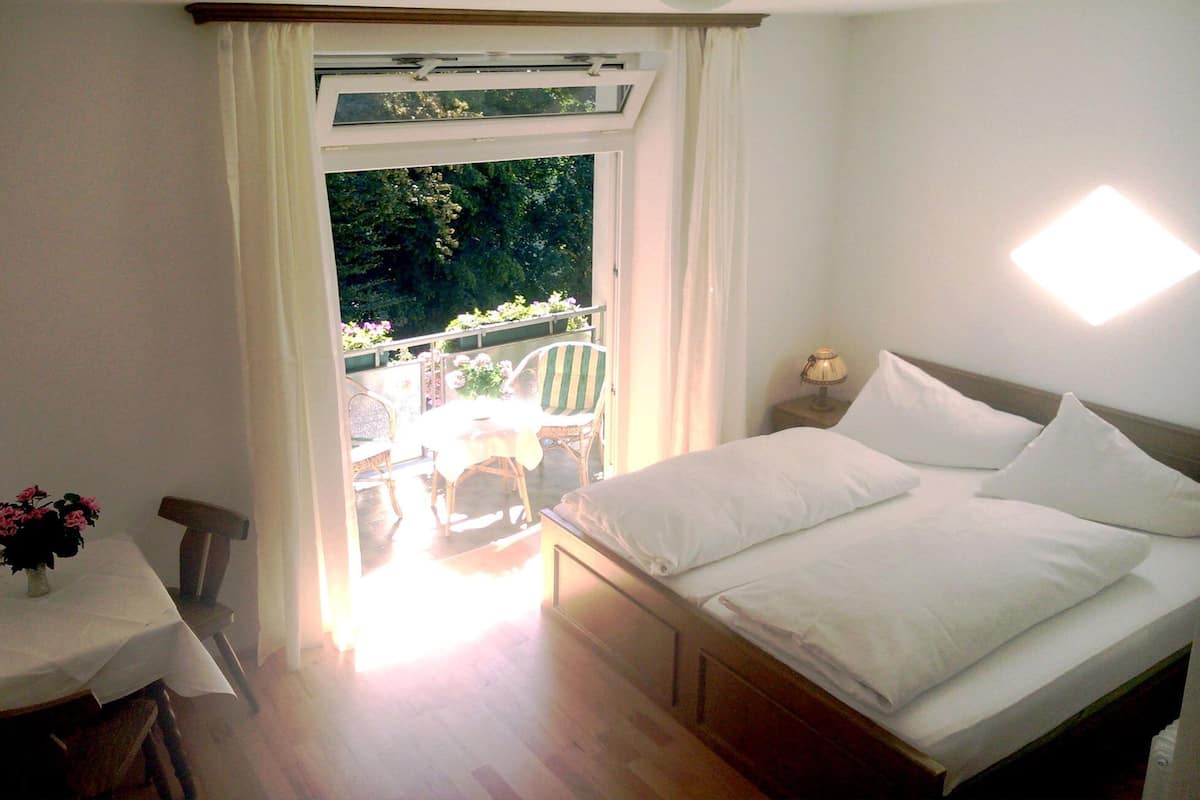 Ein Schlafzimmer mit einem Bett, einem Tisch und einem Stuhl. Ein Fenster mit Vorhängen und ein Balkon mit Pflanzen und einem Stuhl sind ebenfalls vorhanden.