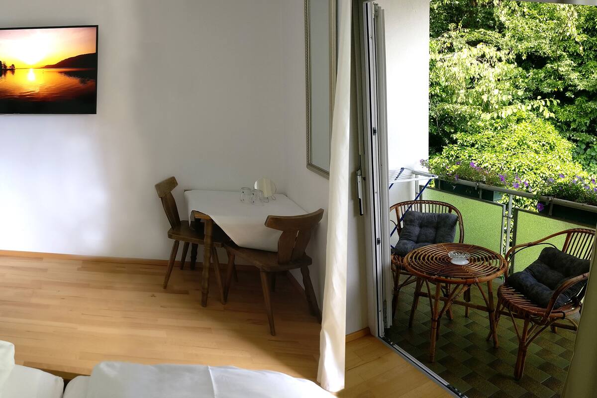 Ein Zimmer mit Bett, Schreibtisch inklusive Computer, Stuhl, Esstisch mit Stühlen sowie ein Balkon mit Korbmöbeln.