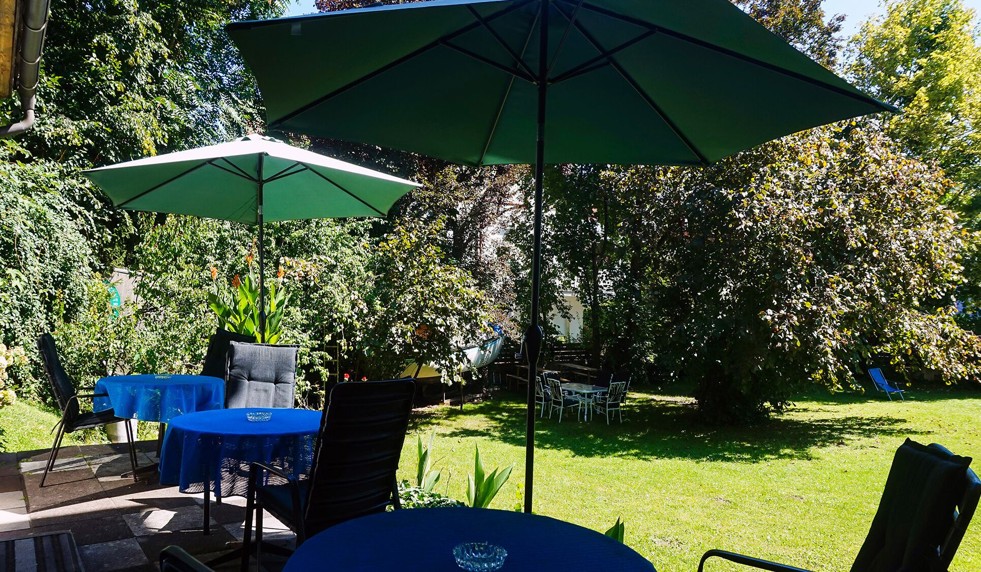 Un espace extérieur aménagé en terrasse, avec des tables et des chaises, protégé par des parasols verts et entouré d’une végétation luxuriante.