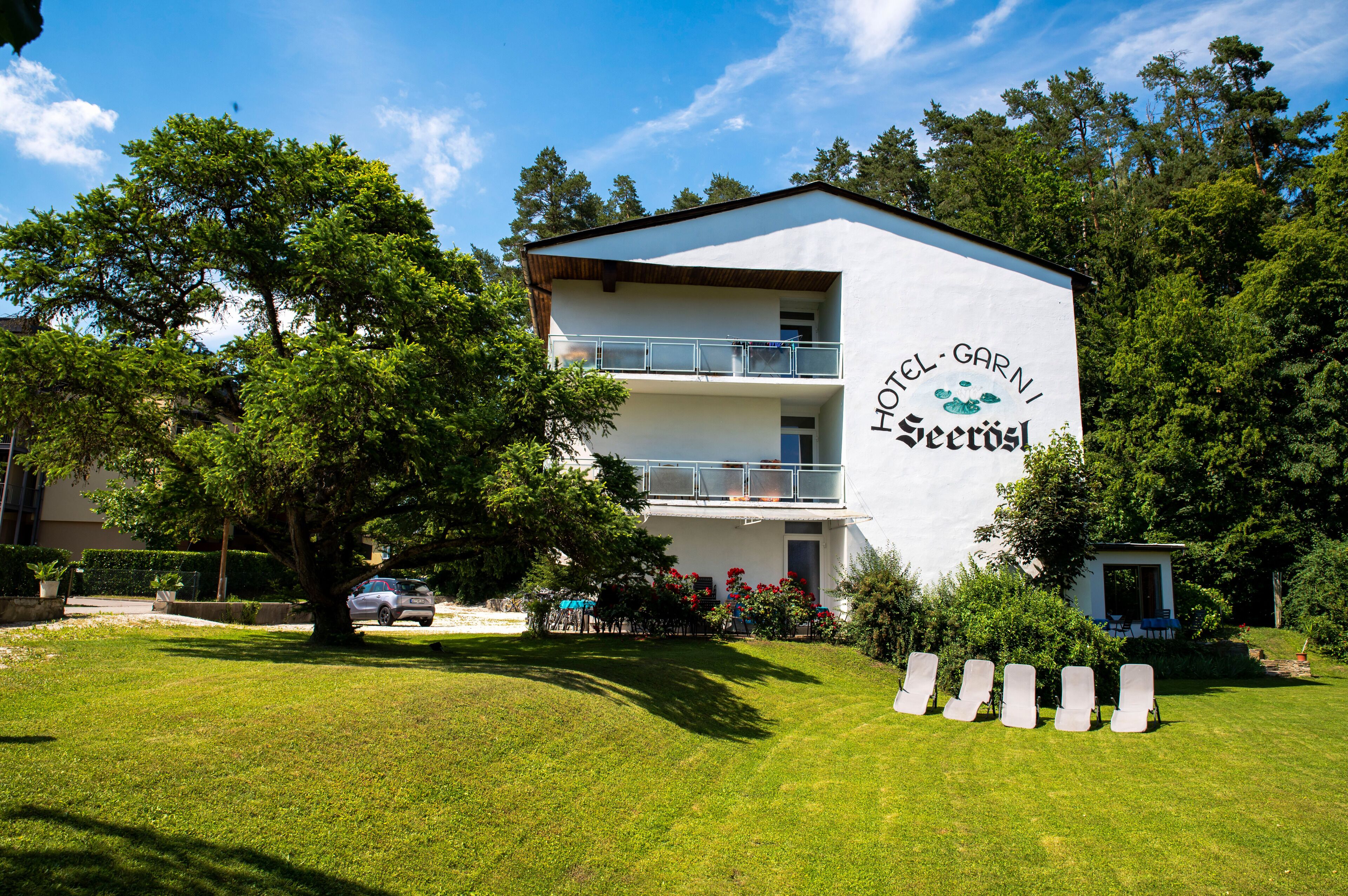 Hotelli nimeltä »Hotel Garni», jonka julkisivu on valkoinen, siinä on parvekkeet, ja se on vehreän kasvillisuuden ympäröimä.