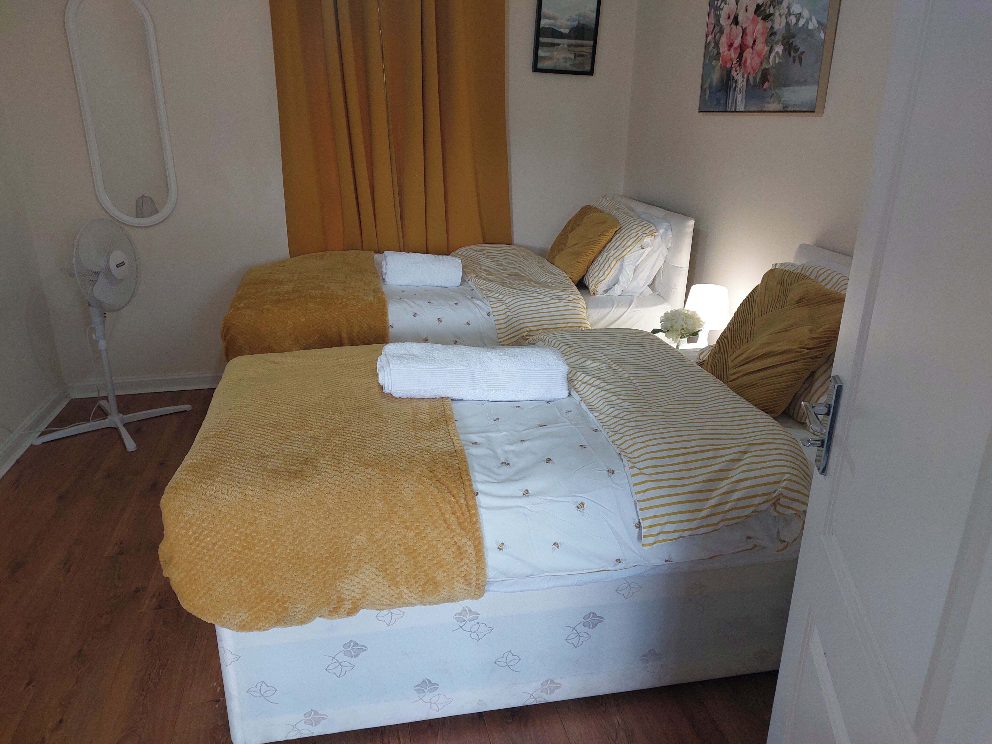 Un lit double avec une couvre-lit blanche à rayures, un plaid marron et un ventilateur sur pied.