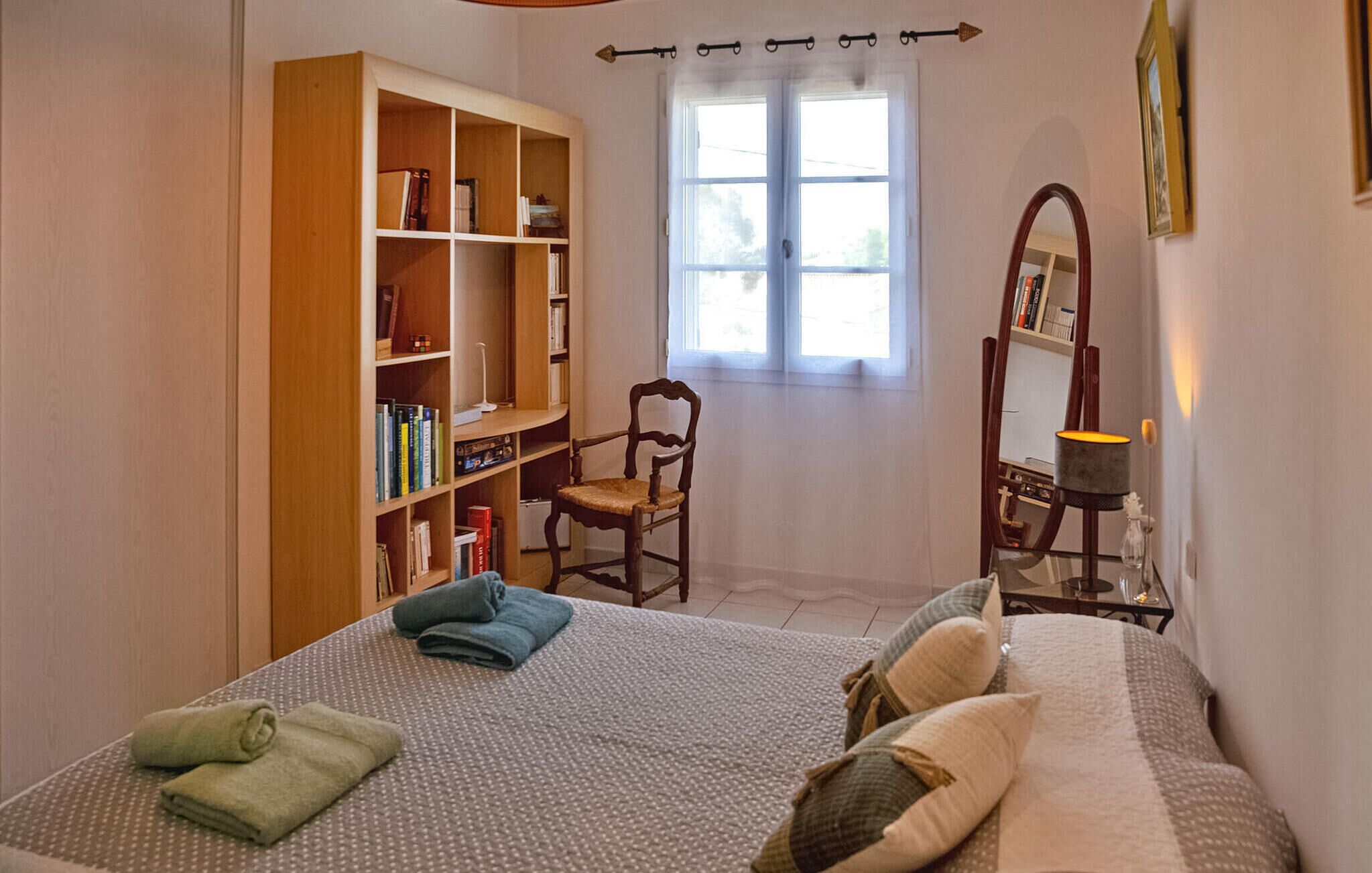 Um quarto com cama, estante de madeira, cadeira, espelho e janela.