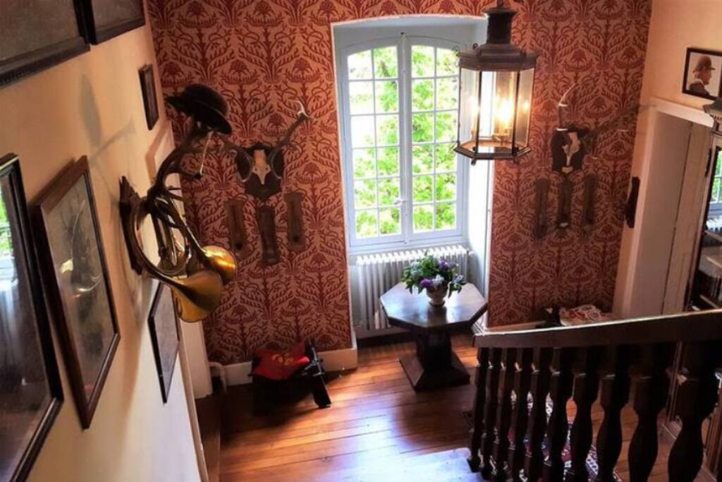 A hallway with patterned wallpaper, a wooden floor, a window with a view of greenery, and a table with a vase of flowers.