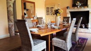 A dining area with a wooden table set for four, surrounded by wicker chairs. A vase with flowers and a fireplace are visible in the background.