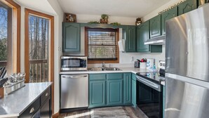 A modern kitchen with teal cabinets, stainless steel appliances, and a window with a view of trees.