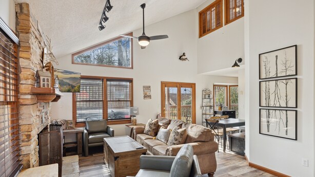 A spacious living room with a large sofa, a wooden coffee table, and a fireplace. There are multiple windows providing natural light and a view of the outdoors.