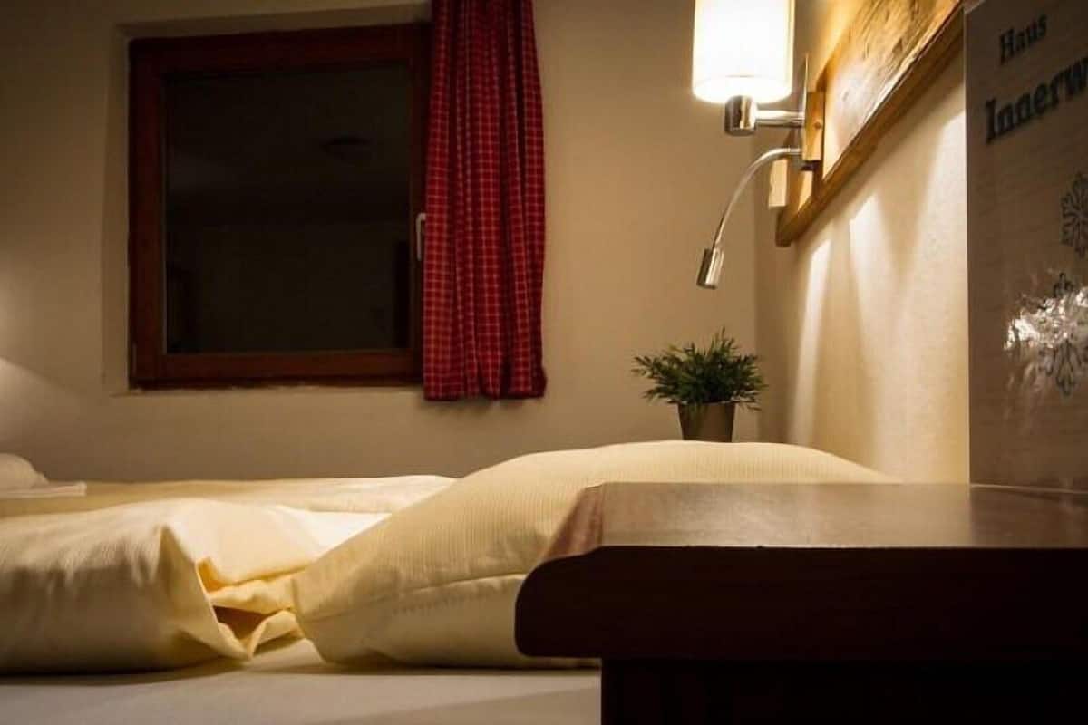 A hotel room with a bed, a small table, a window with a red curtain, and a wall-mounted light fixture.
