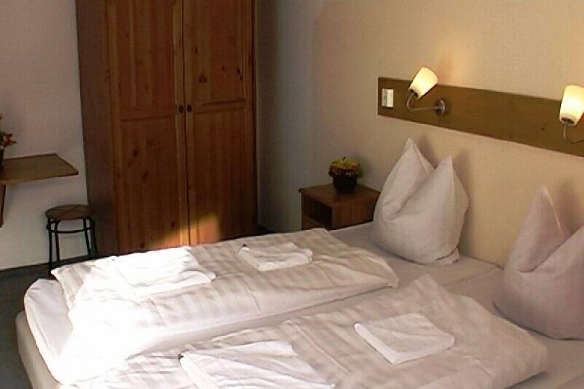 A hotel room with two beds, wooden wardrobe, bedside table with lamp, and a small chair.