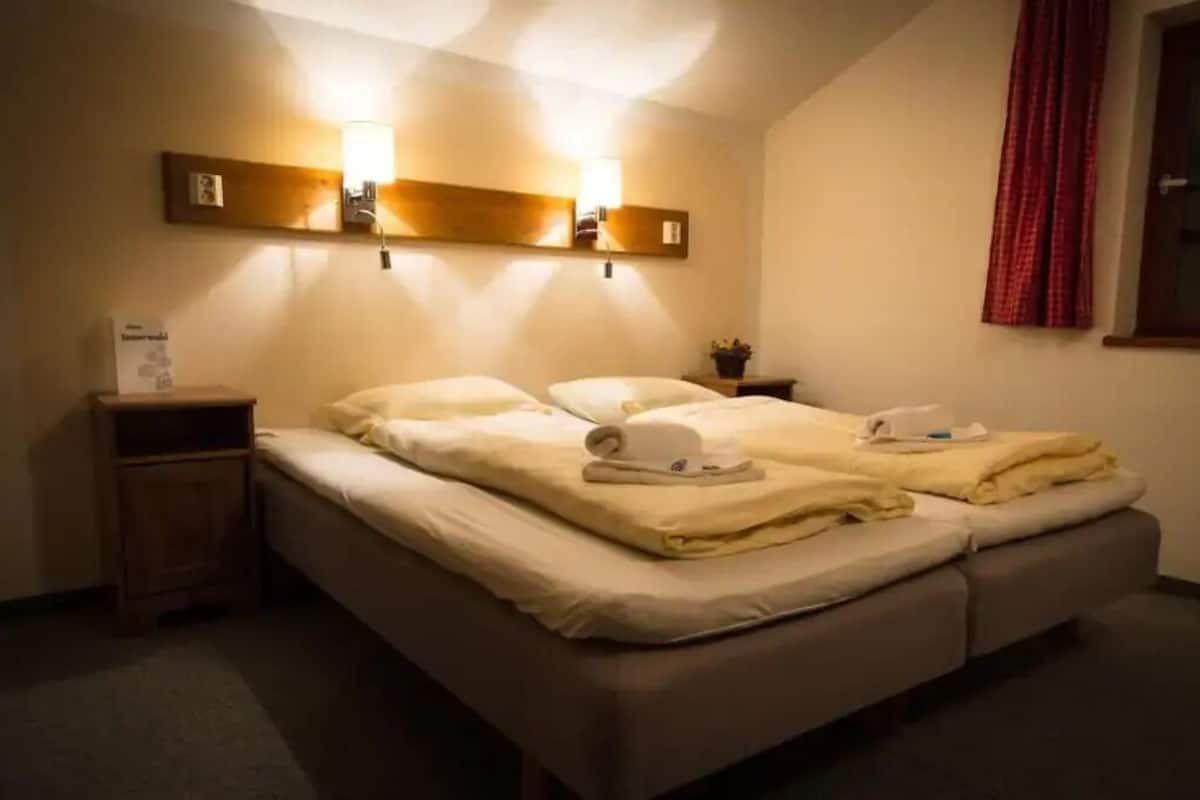 A double bed with two pillows and a blanket, a bedside table, a wall-mounted light fixture, and a window with a red curtain.