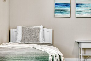 Un lit avec une tête de lit blanche, un oreiller à texture et une couverture verte. Deux tableaux représentant une scène de plage sont accrochés au mur.