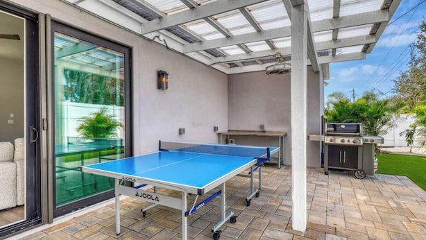 En uteplats med ett bordtennisbord, en grill och en glasdörr som leder in till ett rum.