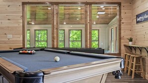 A pool table with balls, a pool cue, and a cue ball in a room with wooden walls and large windows.
