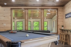 Une table de billard avec des billes, une queue de billard et une bille blanche, dans une pièce aux murs en bois et aux grandes fenêtres.