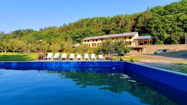 A swimming pool with lounge chairs, a building with a sloped roof, and a forested hillside in the background. - Restland Dilijan Family Hotel  (Dilijan)