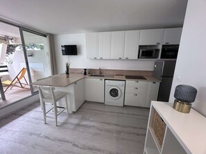 A compact kitchen with white cabinetry, a washing machine, a microwave, and a dining area with a chair. - 2-room apartment with pool, tennis and private parking 50m from the beach (Port Camargue)