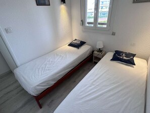 A small, simple room with two single beds, a nightstand with a lamp, and a window with a view of the outside. - 2-room apartment with pool, tennis and private parking 50m from the beach (Port Camargue)
