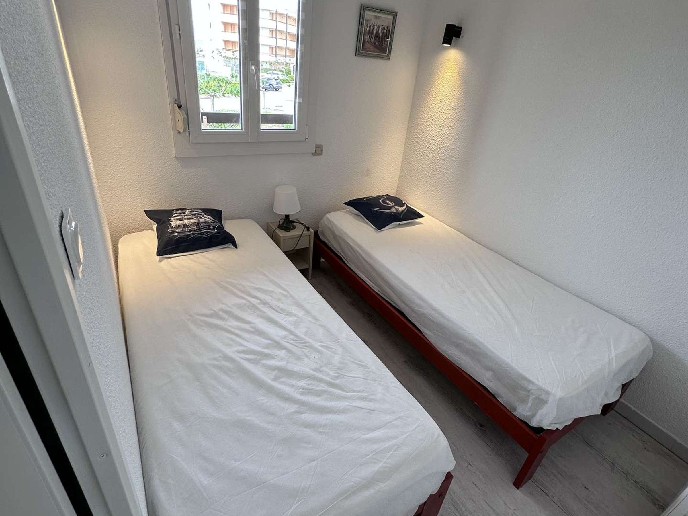 A room with two single beds, a nightstand with a lamp, a window with blinds, and a wall-mounted light fixture.